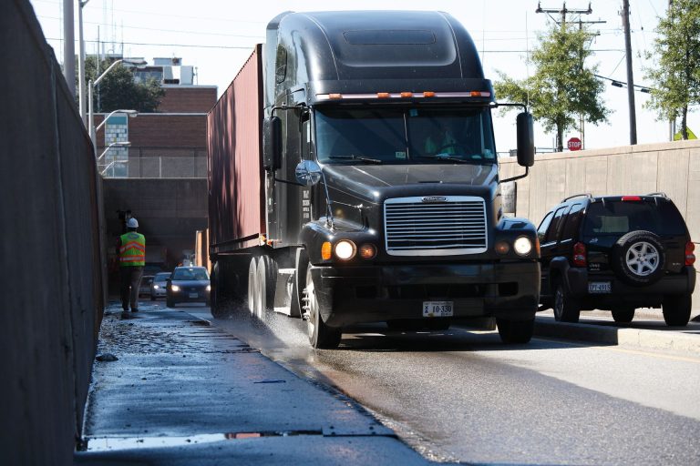 A dark tractor-trailer drives along an urban street as a worker stands beside the road