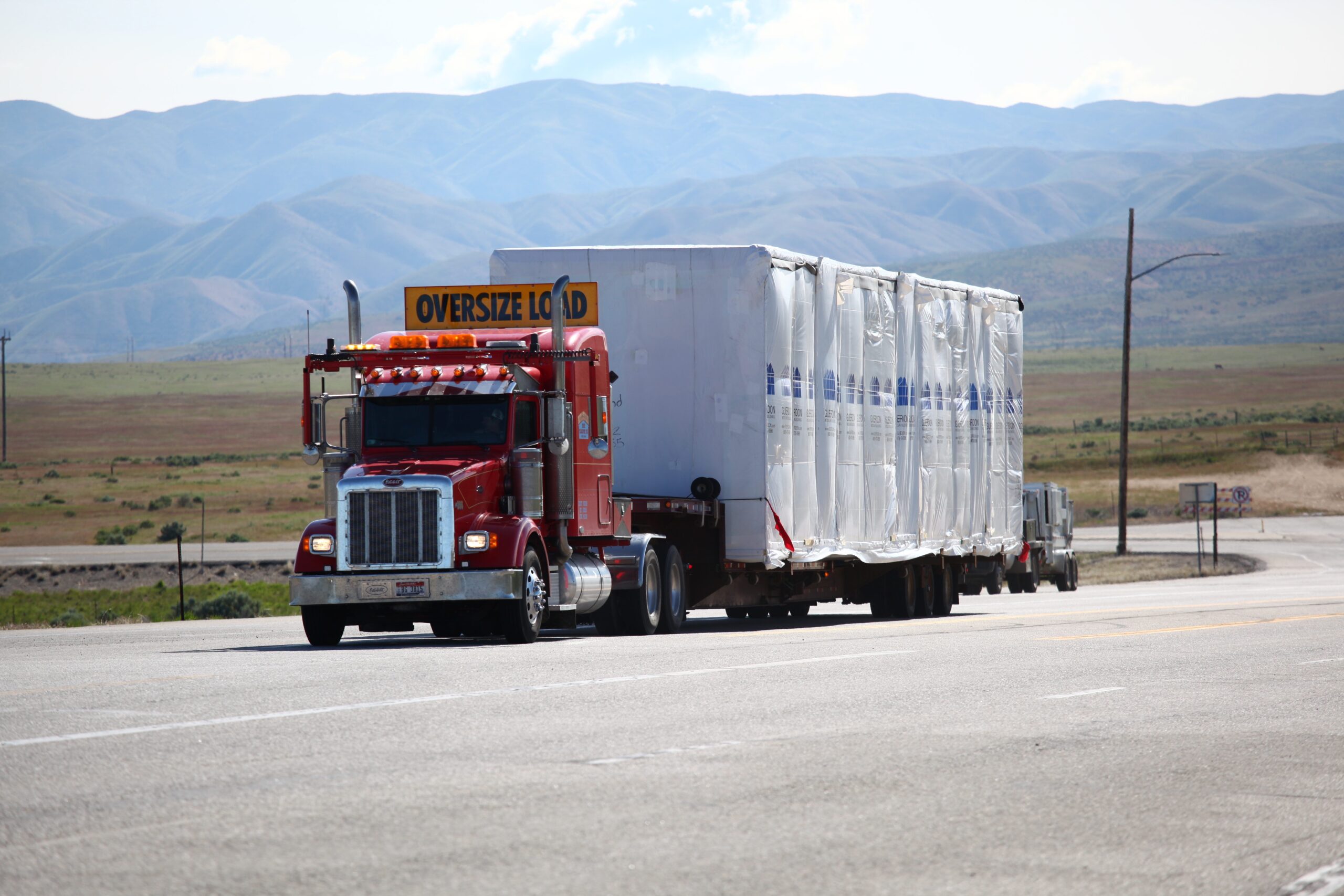 A red semi truck hauling an oversized load along a road with mountains in the background