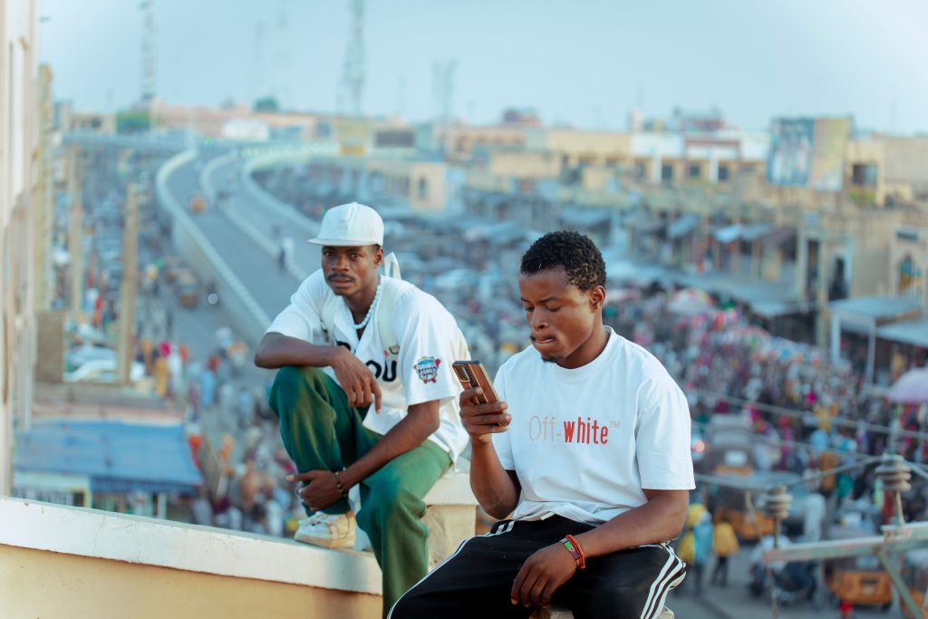 Two young men sit on a rooftop overlooking a bustling city market, feeling the pulse of urban life